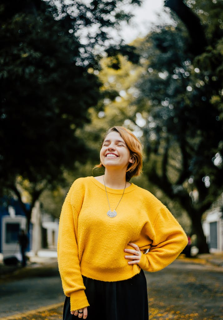 Adult woman in a yellow sweater standing outdoors, smiling with eyes closed, embodying fashion and joy.